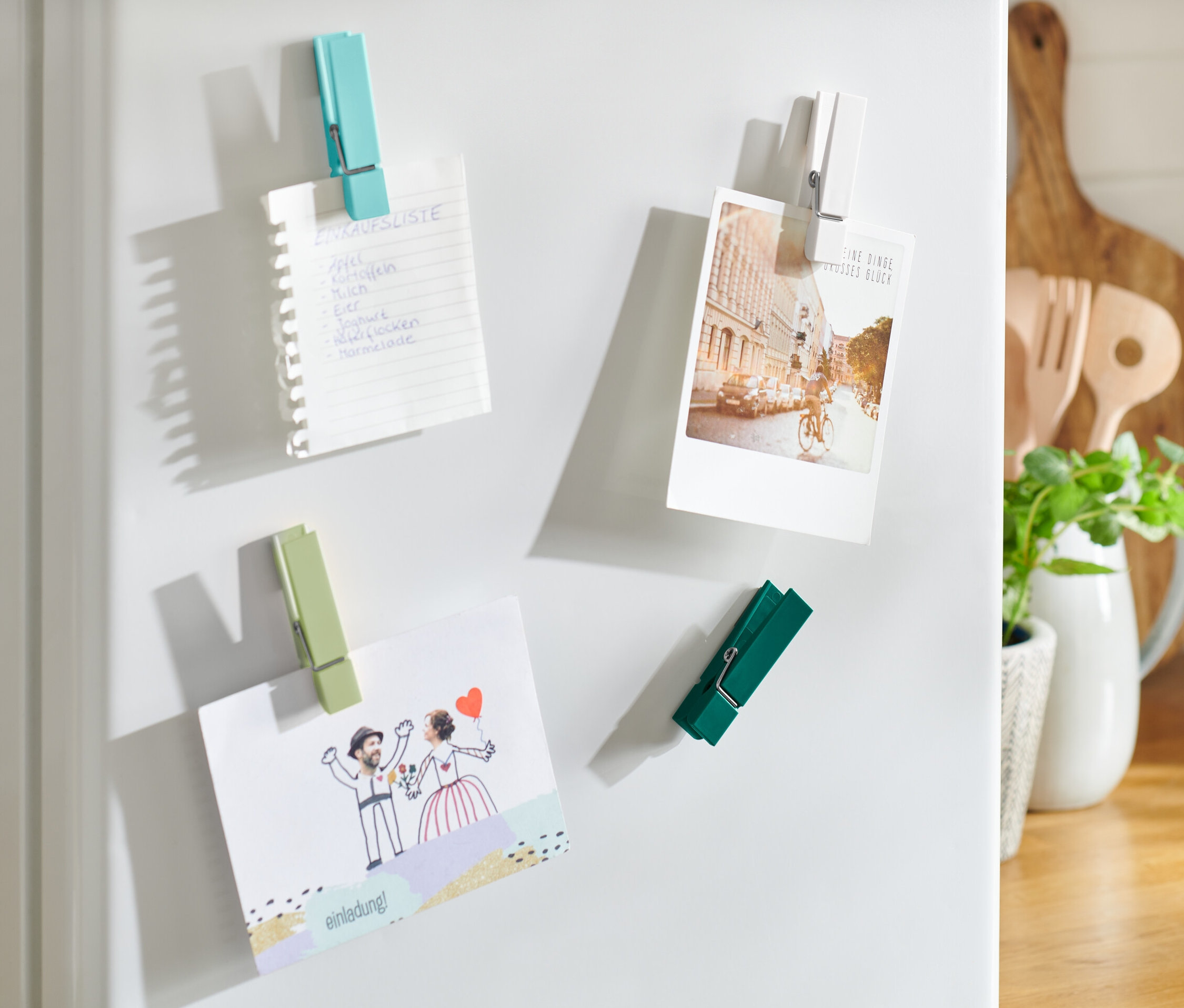 Čtyři magnety na lednici drží nákupní seznam, pohlednici a pozvánku.