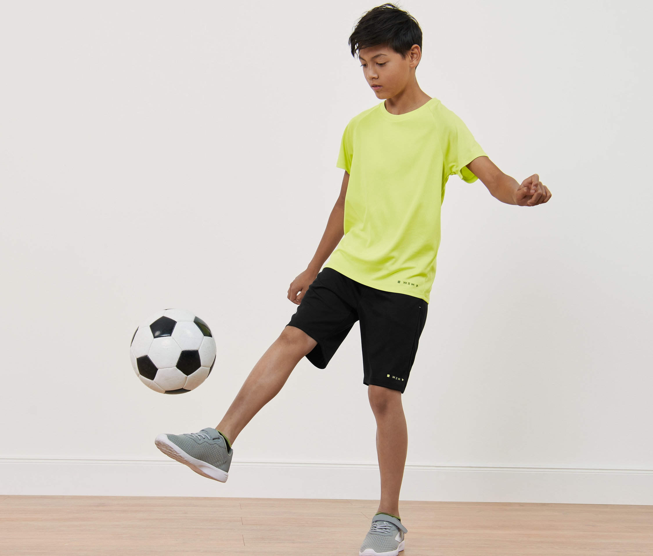 Chlapec kope do fotbalového míče. Má na sobě černé sportovní šortky a neonově žluté funkční tričko. Dětská funkční trička 2 ks.