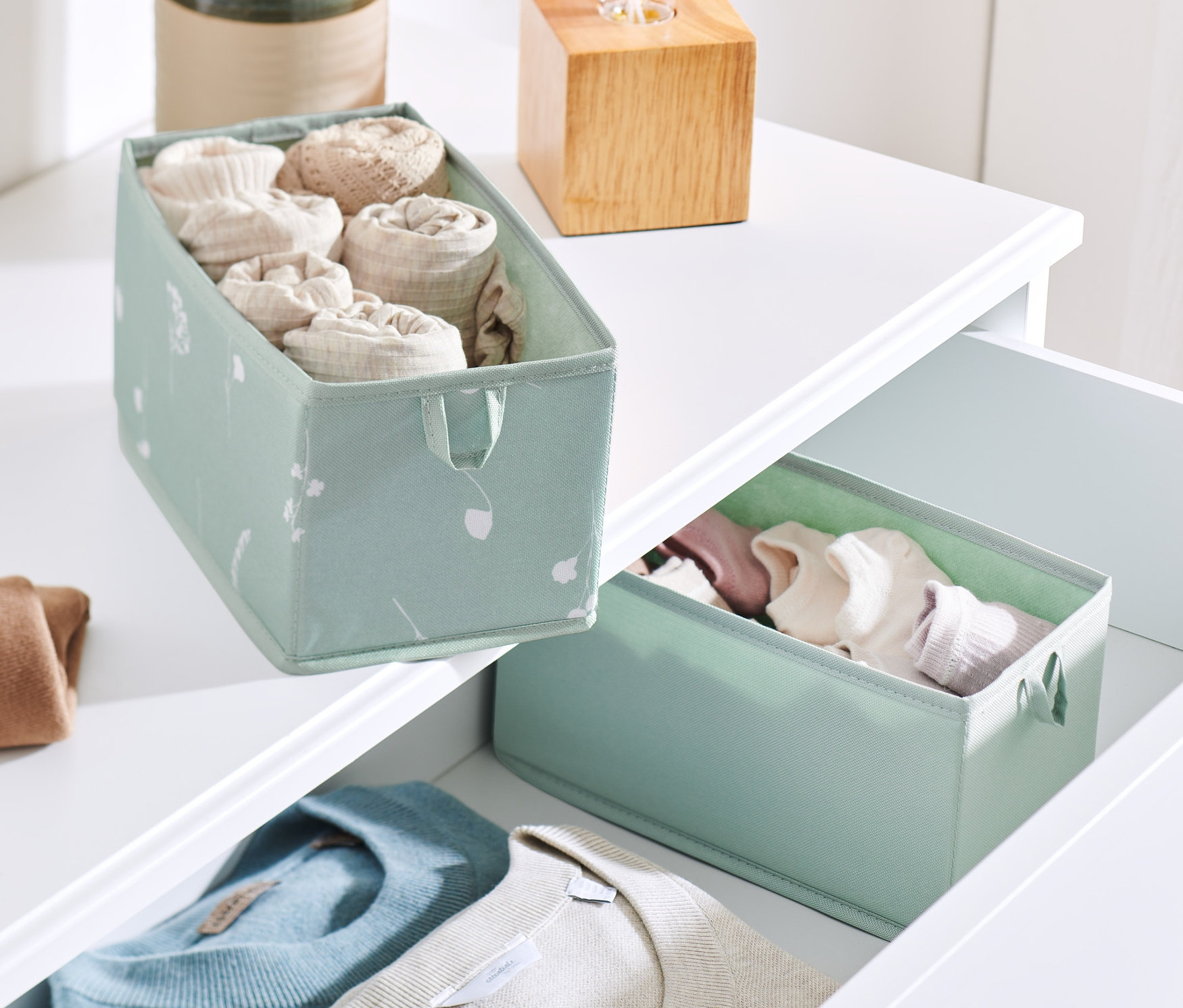 Dva střední úložné boxy do zásuvky na bílém prádelníku. Jeden box je na horní části prádelníku a obsahuje stočené oblečení. Druhý je v otevřené zásuvce s oblečením.