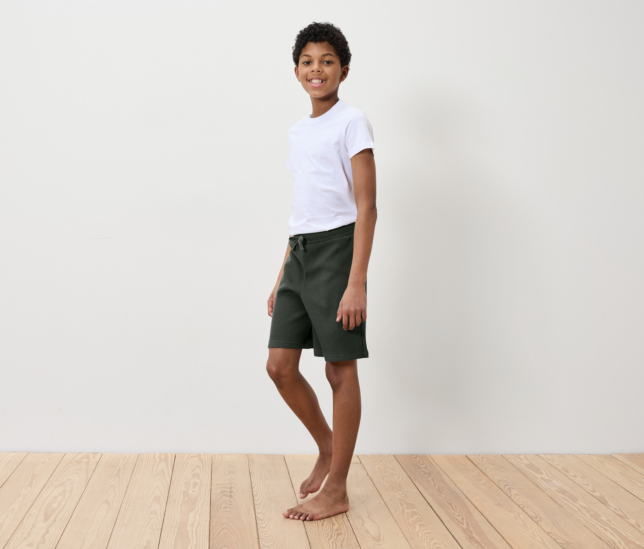 Chlapec v bílém tričku a zelených šortkách stojí na dřevěné podlaze před bílou zdí.