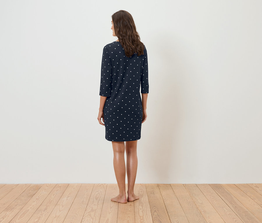 Žena stojí zády k fotoaparátu a má na sobě tmavě modré šaty s bílými puntíky.