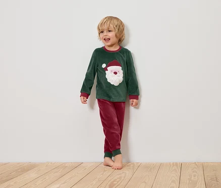 Chlapec v zeleném vánočním pyžamu s obrázkem Santa Clause stojí na dřevěné podlaze před bílou stěnou.