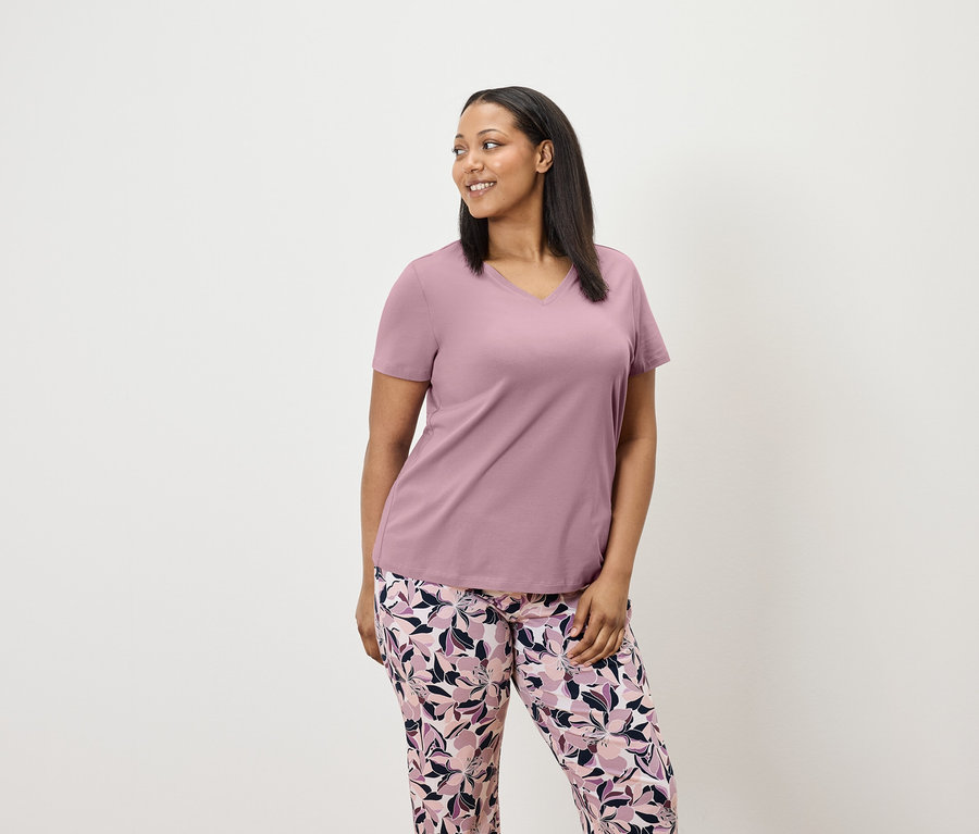 Žena v lila tričku s krátkým rukávem a květinových kalhotách se dívá vpravo.