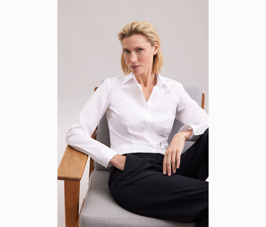 Žena sedí na židli a má na sobě bílou halenku Seidensticker Slim Fit non iron.