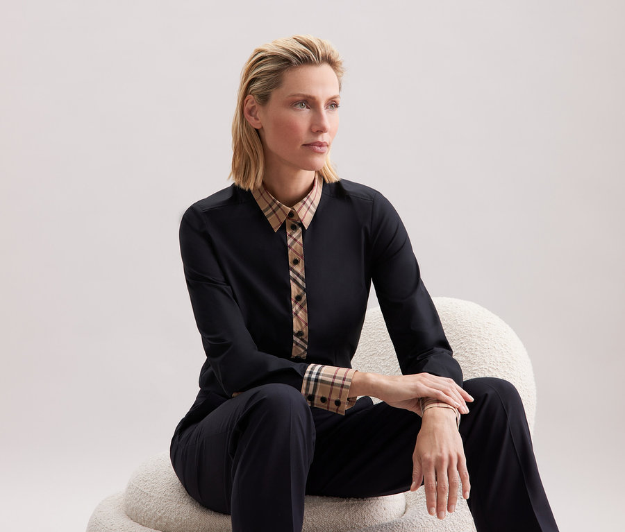 Blondýna sedí na bílé židli v černém oblečení s károvanými detaily.