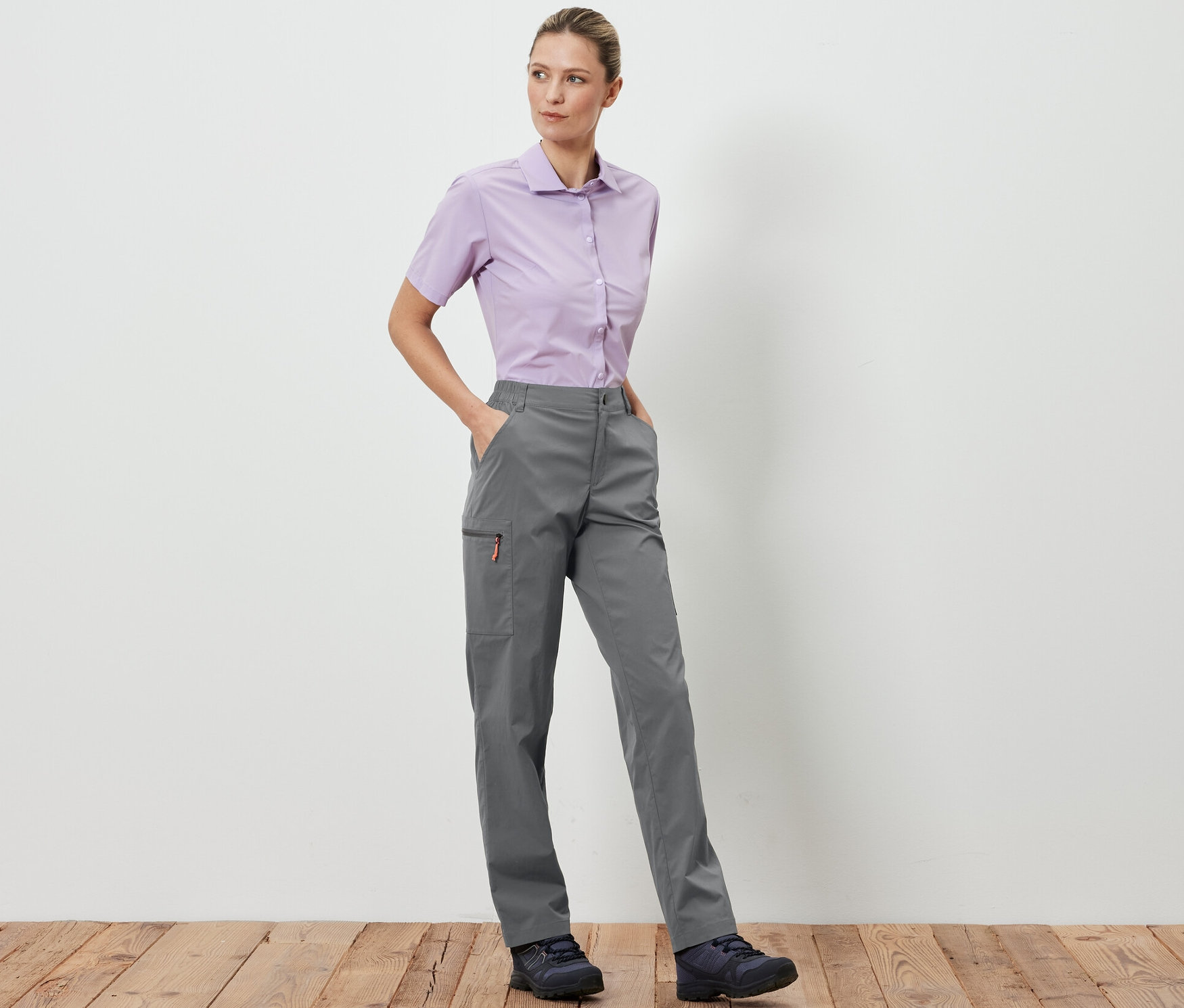 Žena stojí a pózuje v šedých outdoorových kalhotách Sbalitelné outdoorové kalhoty a světle fialové košili.