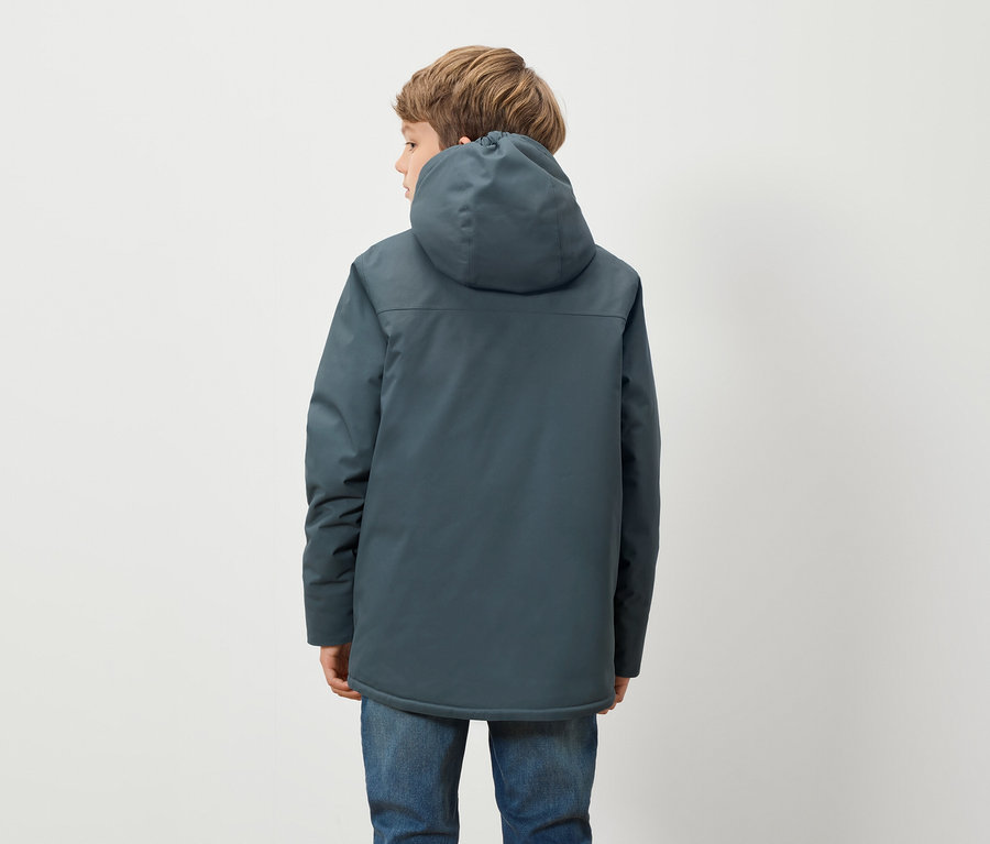 Chlapec v modré bundě s kapucí a modrých džínách stojí zády k fotoaparátu před bílým pozadím.