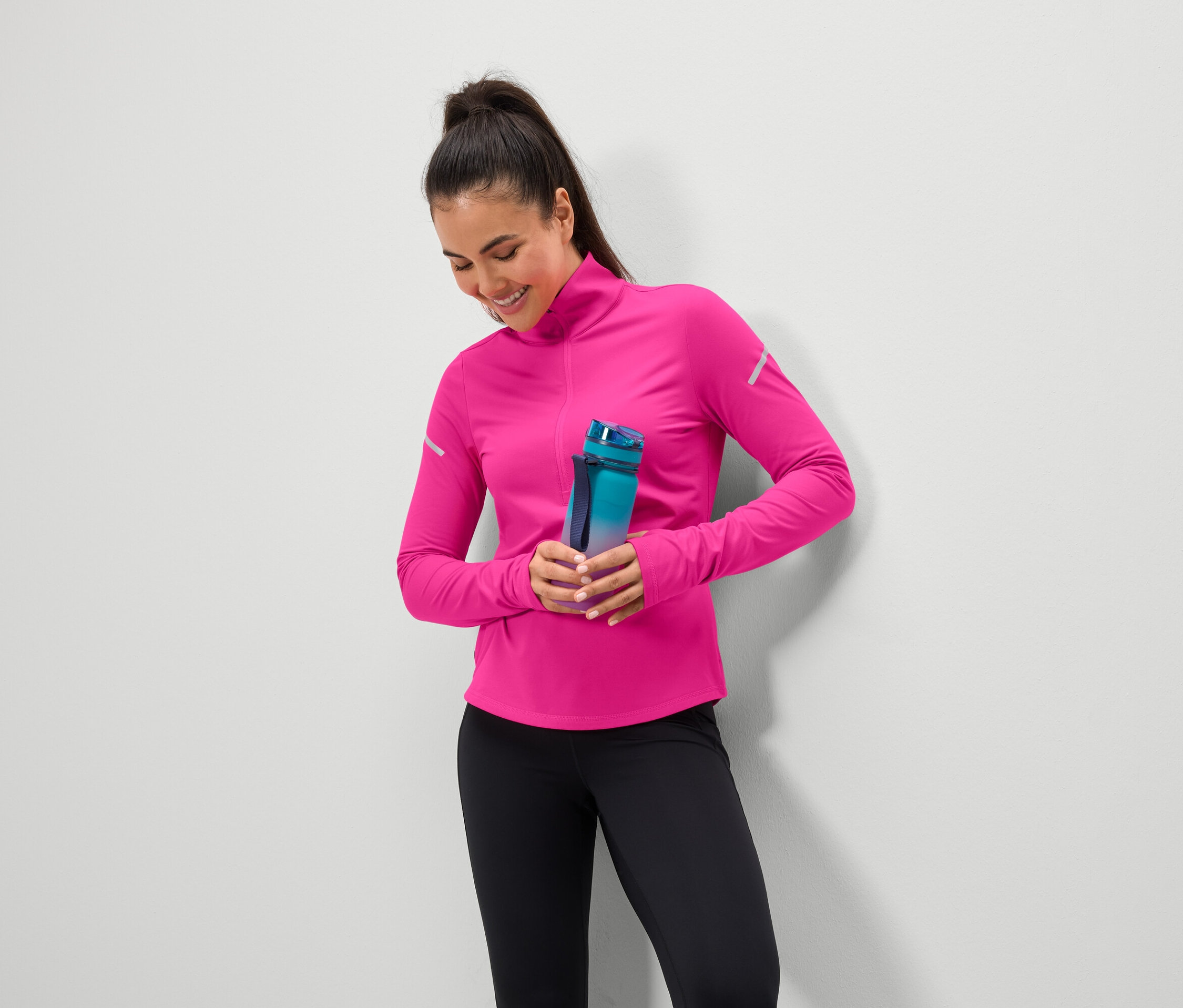 Usmívající se žena drží modrozelenou láhev na vodu, má na sobě růžový top a černé legíny.