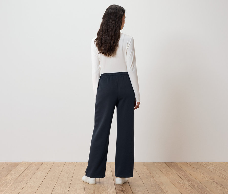 Žena s dlouhými vlnitými vlasy stojí zády a má na sobě bílý top s dlouhým rukávem a tmavě modré široké kalhoty.