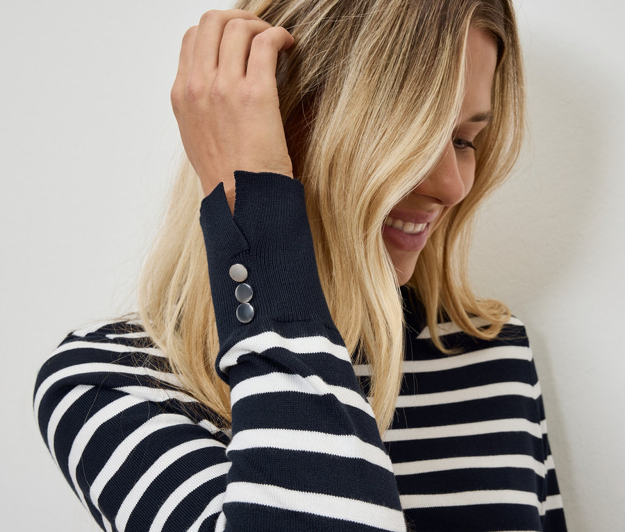 Blondýna si rukou upravuje vlasy, oblečená v pruhovaném svetru s knoflíky na rukávu.