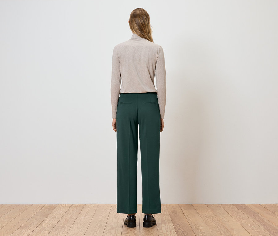 Žena stojí zády, má na sobě béžový rolák a zelené kalhoty.