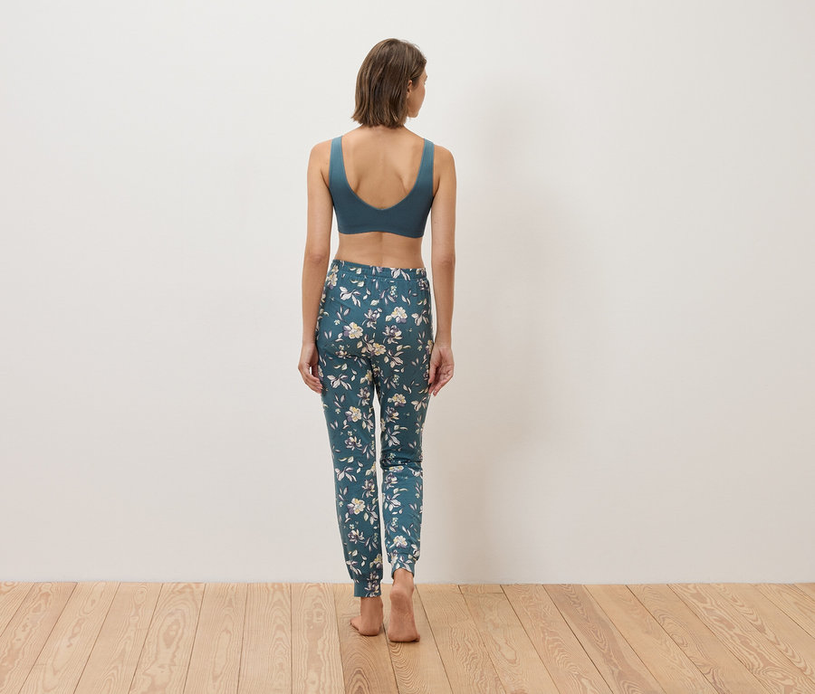 Pohled zezadu na ženu v modré sportovní podprsence a květinových kalhotách na dřevěné podlaze.