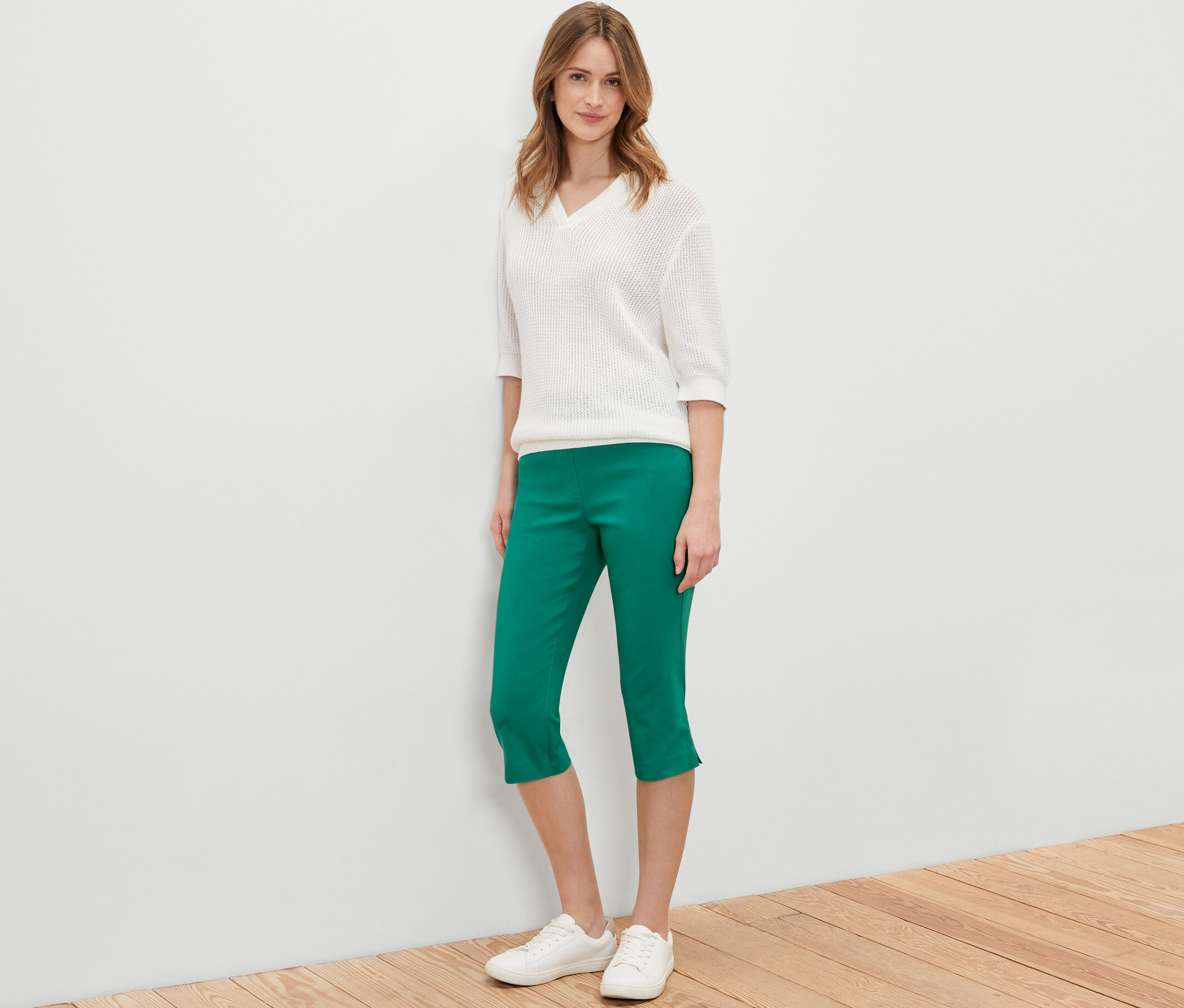 Žena stojí a pózuje v zelených 3/4 bengalínových kalhotách, bílém svetru a bílých botách.