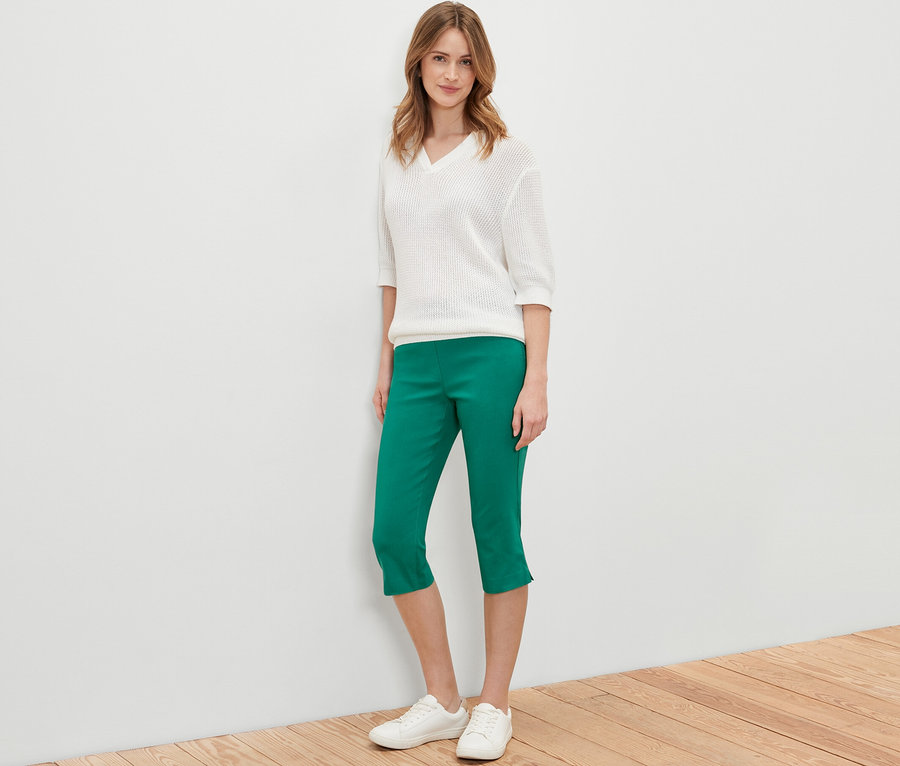 Žena stojí a pózuje v zelených 3/4 bengalínových kalhotách, bílém svetru a bílých botách.