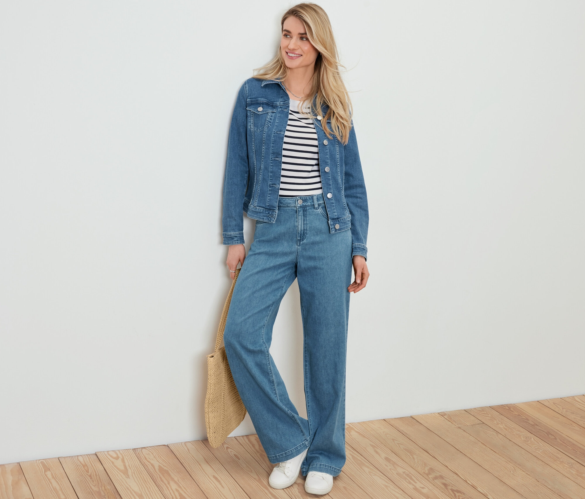 Blondýna opřená o bílou zeď má na sobě džínovou bundu, pruhovaný top, džíny se širokými nohavicemi a drží kabelku lýkového vzhledu.