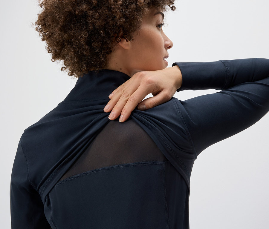 Žena si přetahuje funkční bundu přes rameno a ukazuje síťovaný panel na zádech.