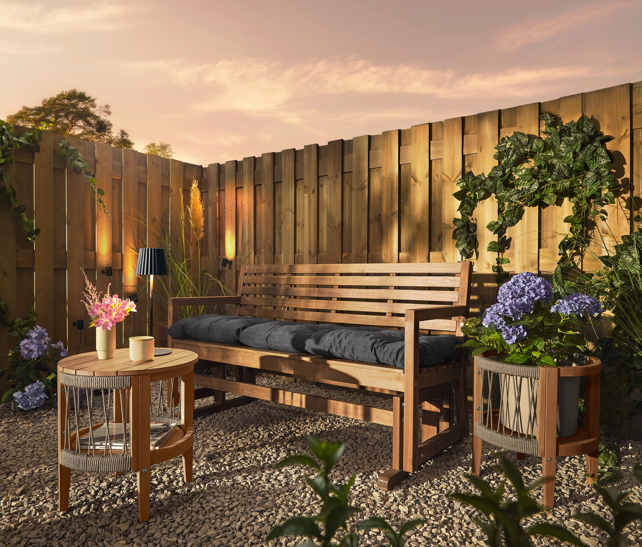 Dřevěná lavice s polštáři stojí na štěrkovém podkladu před dřevěným plotem. Na plotě jsou připevněny 3 solární svítidla na okap.