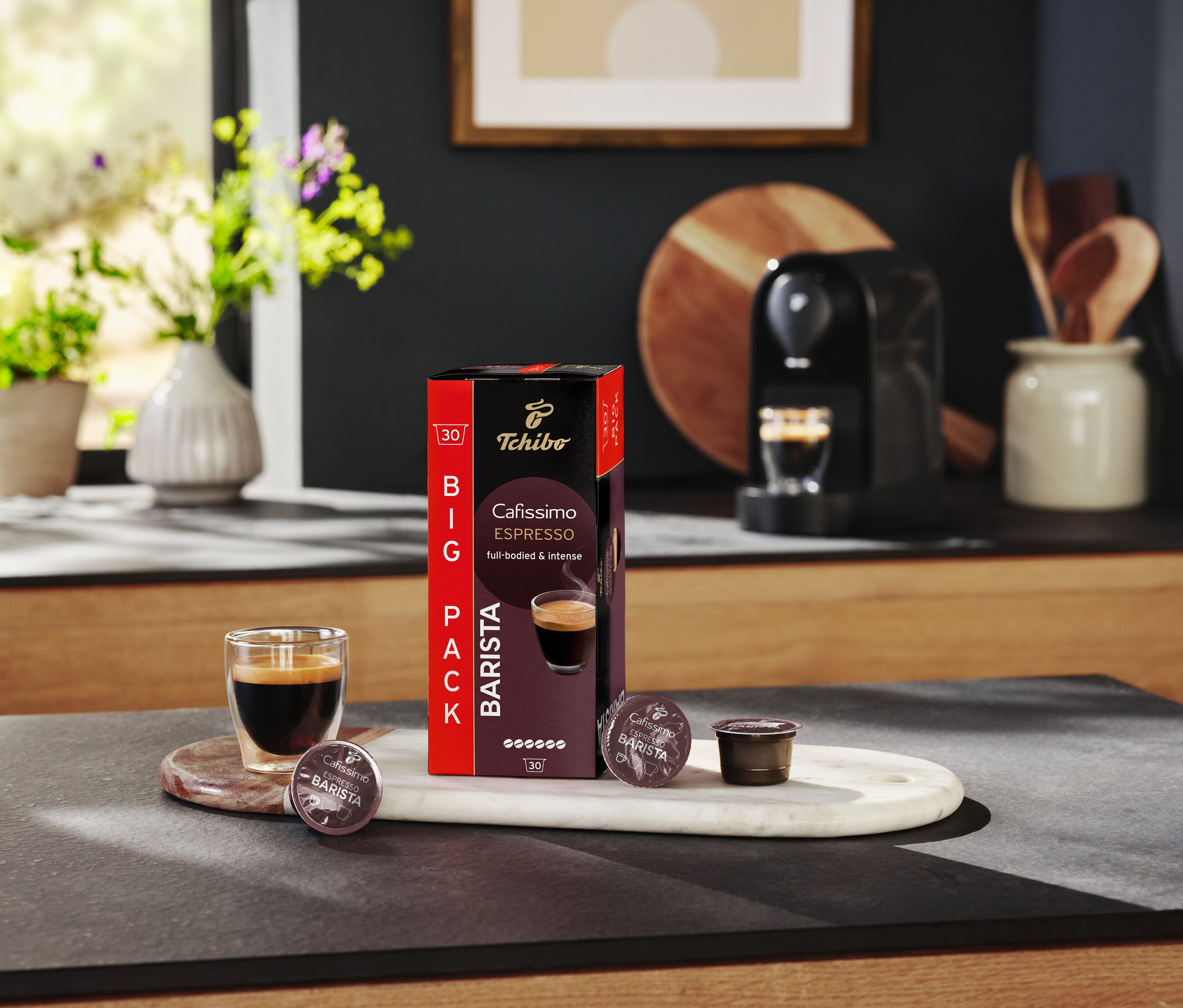 Sklenice espressa stojí vedle balení kapslí Tchibo Cafissimo Barista na kuchyňském stole.