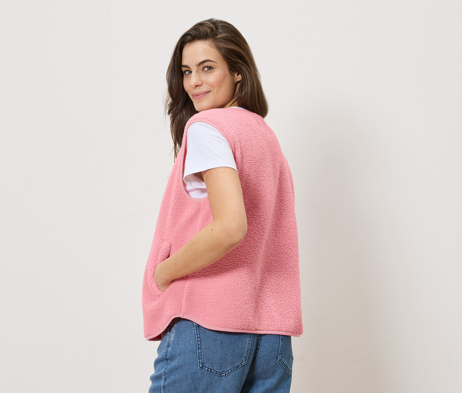 Žena se ohlíží přes rameno a má na sobě plyšovou vestu přes bílé tričko a džíny.