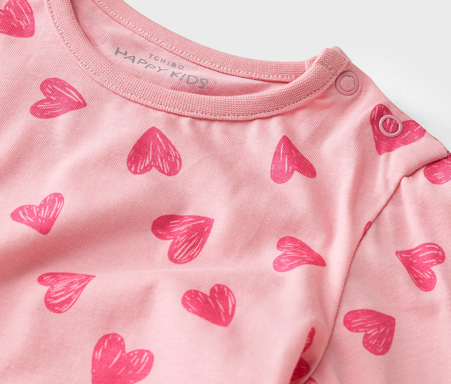 Detail růžového trička s dlouhým rukávem se vzorem srdce a patentkami na límci.