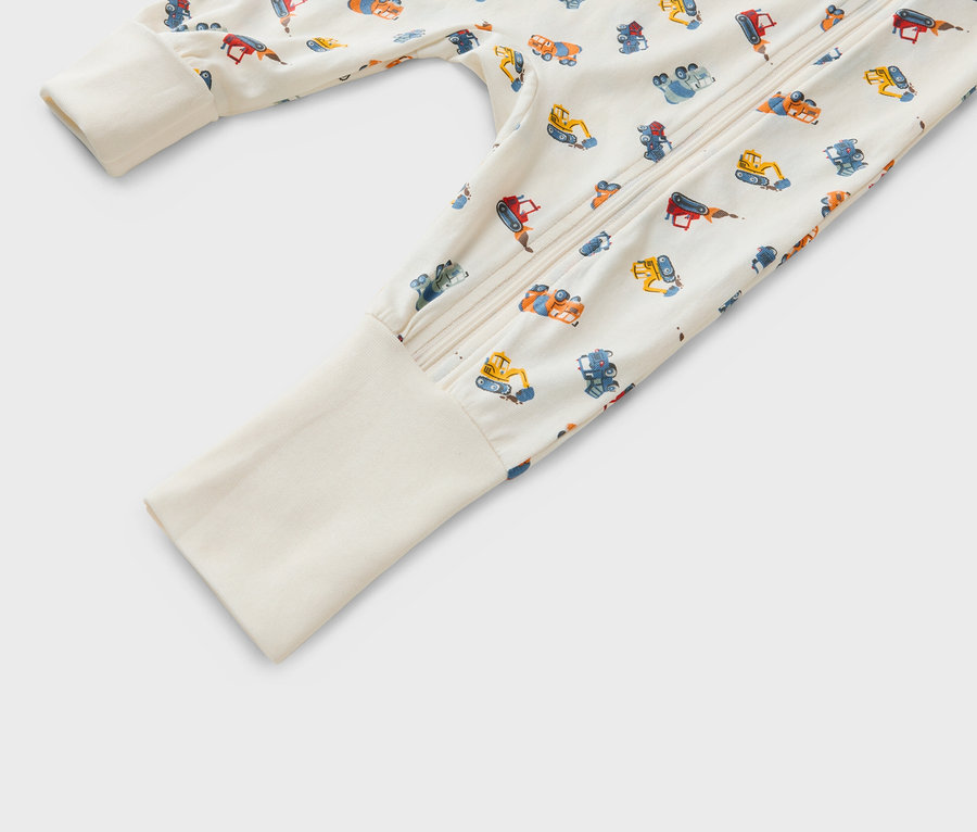 Detailní záběr na dětské pyžamo na zip se vzorem různých stavebních vozidel na béžovém pozadí.