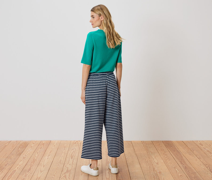 Žena stojí zády ke kameře, má na sobě zelený top a modro-bílé pruhované culottes kalhoty.