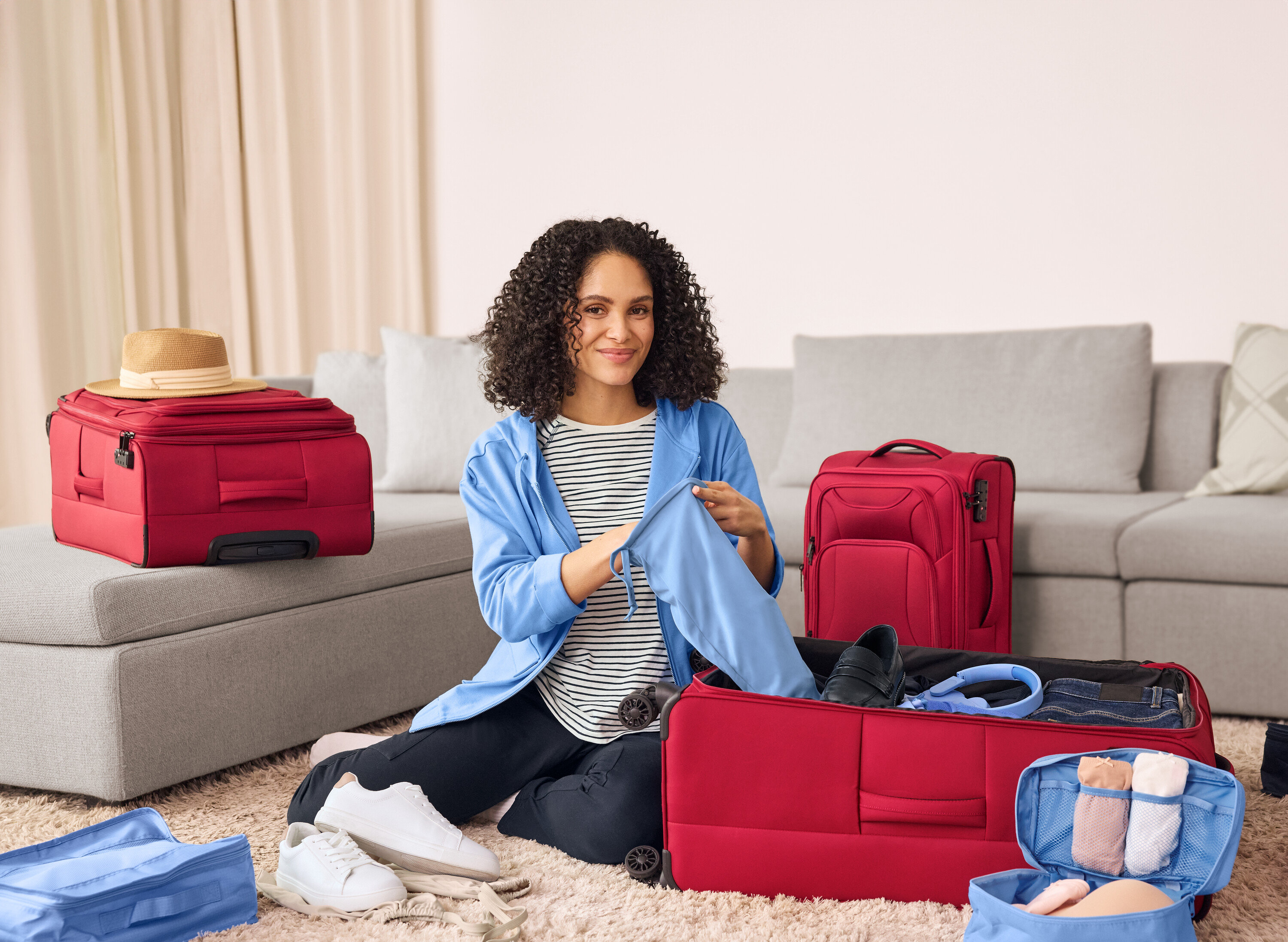 Žena sedí na zemi a balí tmavě modré tepláky s cargo kapsami do červeného kufru. Vedle ní leží kožené tenisky.