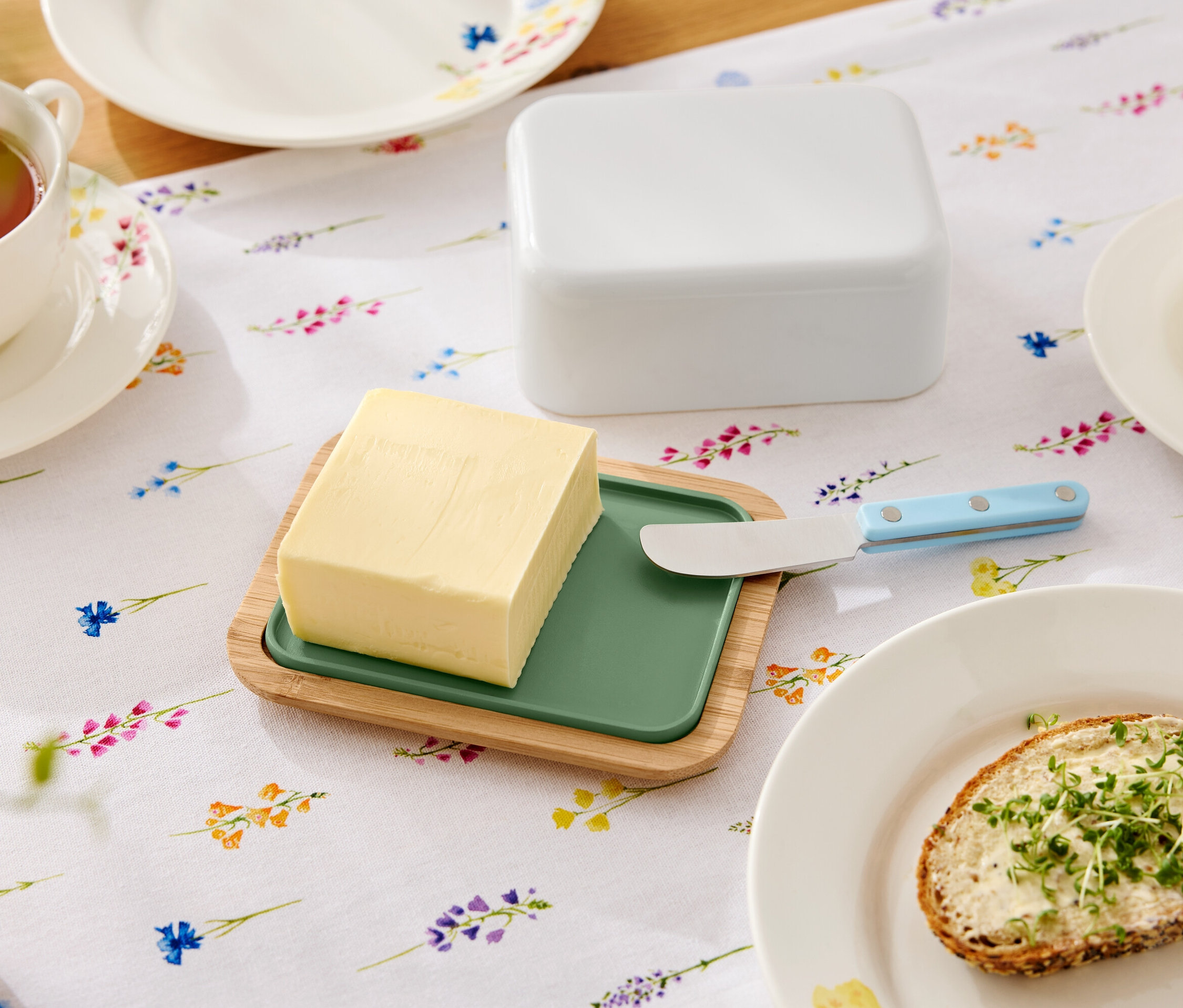 Na květinovém stole leží máslo na dřevěném talíři se zelenou vložkou, nůž na máslo, máslenka, šálek čaje a talíř s chlebíčkem s máslem.