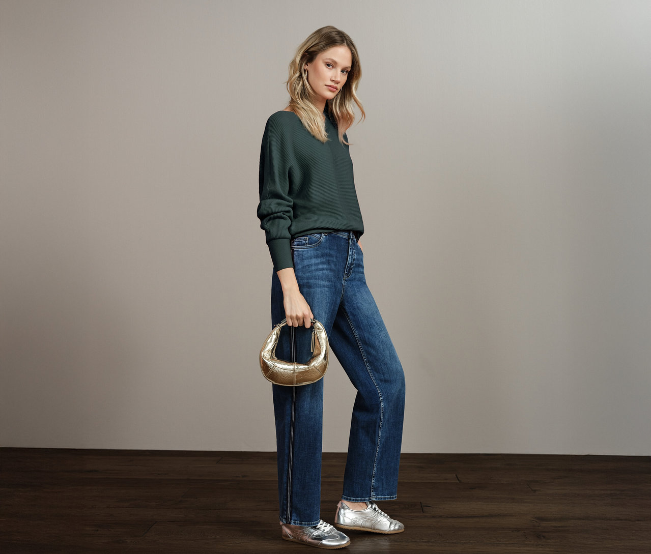 Žena má na sobě strukturovaný svetr Street One, modré džíny, stříbrné boty a zlatou kabelku.