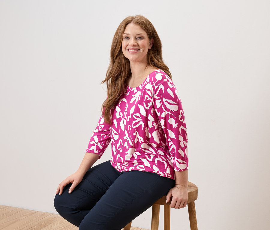 Žena s dlouhými, zrzavými vlasy sedí na dřevěné židli a má na sobě růžovou halenku s bílými listy a tmavě modré kalhoty.