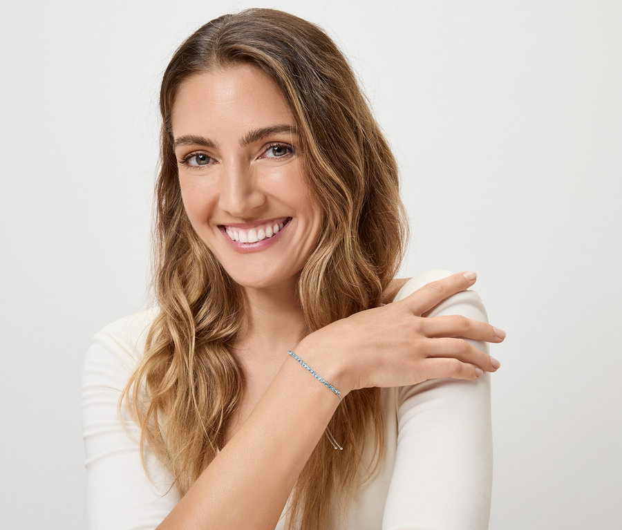 Usmívající se žena s blond vlnitými vlasy a modrým náramkem na levém zápěstí na bílém pozadí.