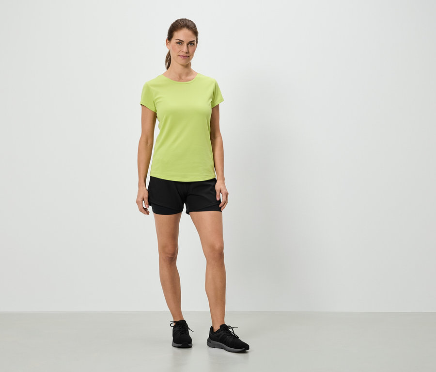 Žena stojí v světle zeleném tričku, černých šortkách a černých teniskách před bílou stěnou.