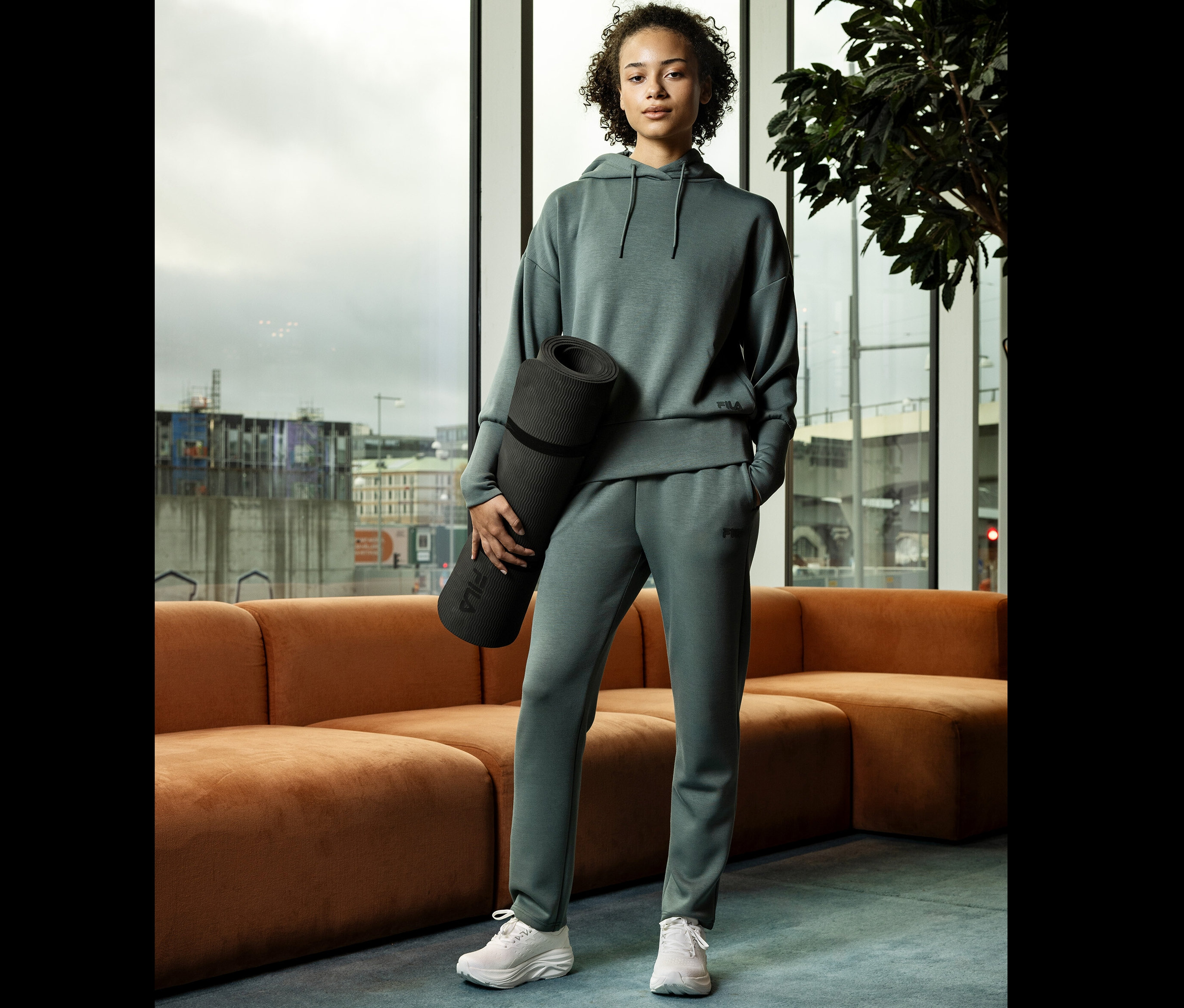 Žena v zelené teplákové soupravě drží podložku na jógu před oknem.