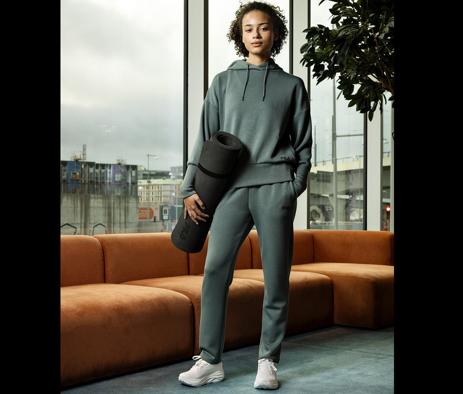 Žena v zelené teplákové soupravě drží podložku na jógu před oknem.