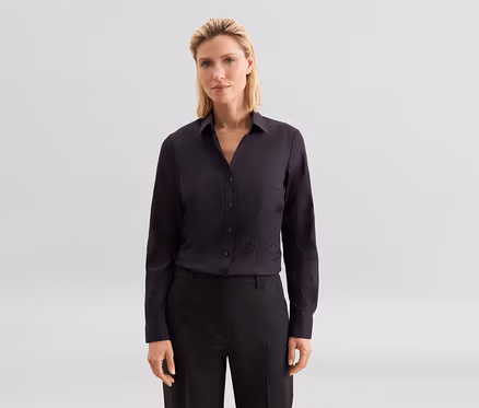 Žena s blond vlasy v tmavé košili s dlouhým rukávem a tmavých kalhotách, dívající se do kamery.