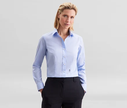 Žena má na sobě světle modrou halenku slim fit non iron Seidensticker a černé kalhoty, ruce v kapsách.