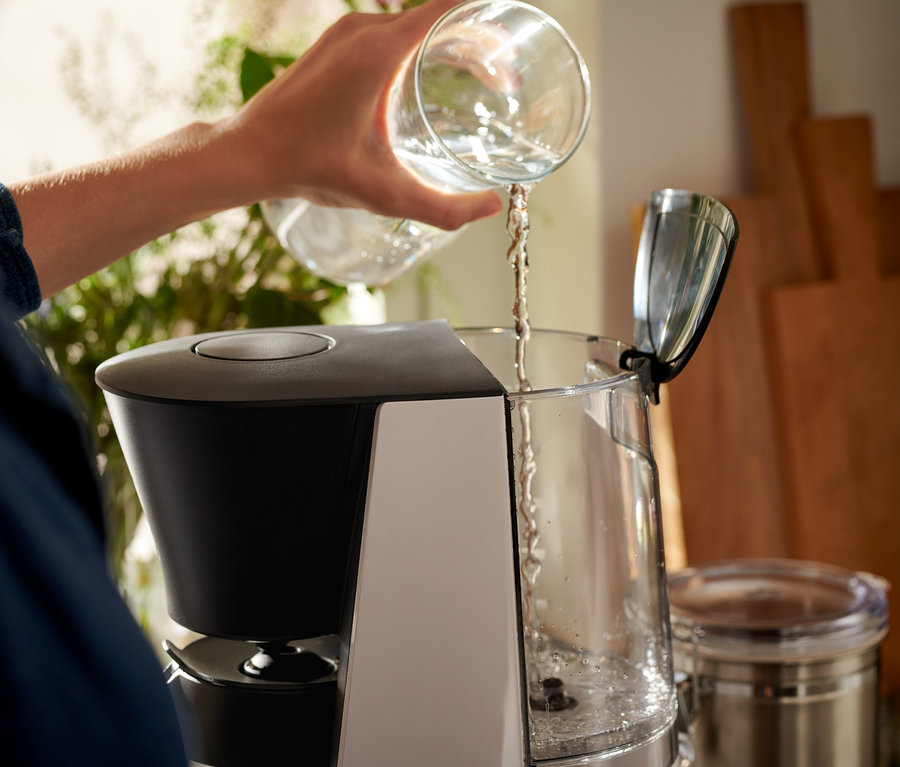 Žena nalévá vodu z čiré sklenice do šedočerného kávovaru »Let's Brew«.