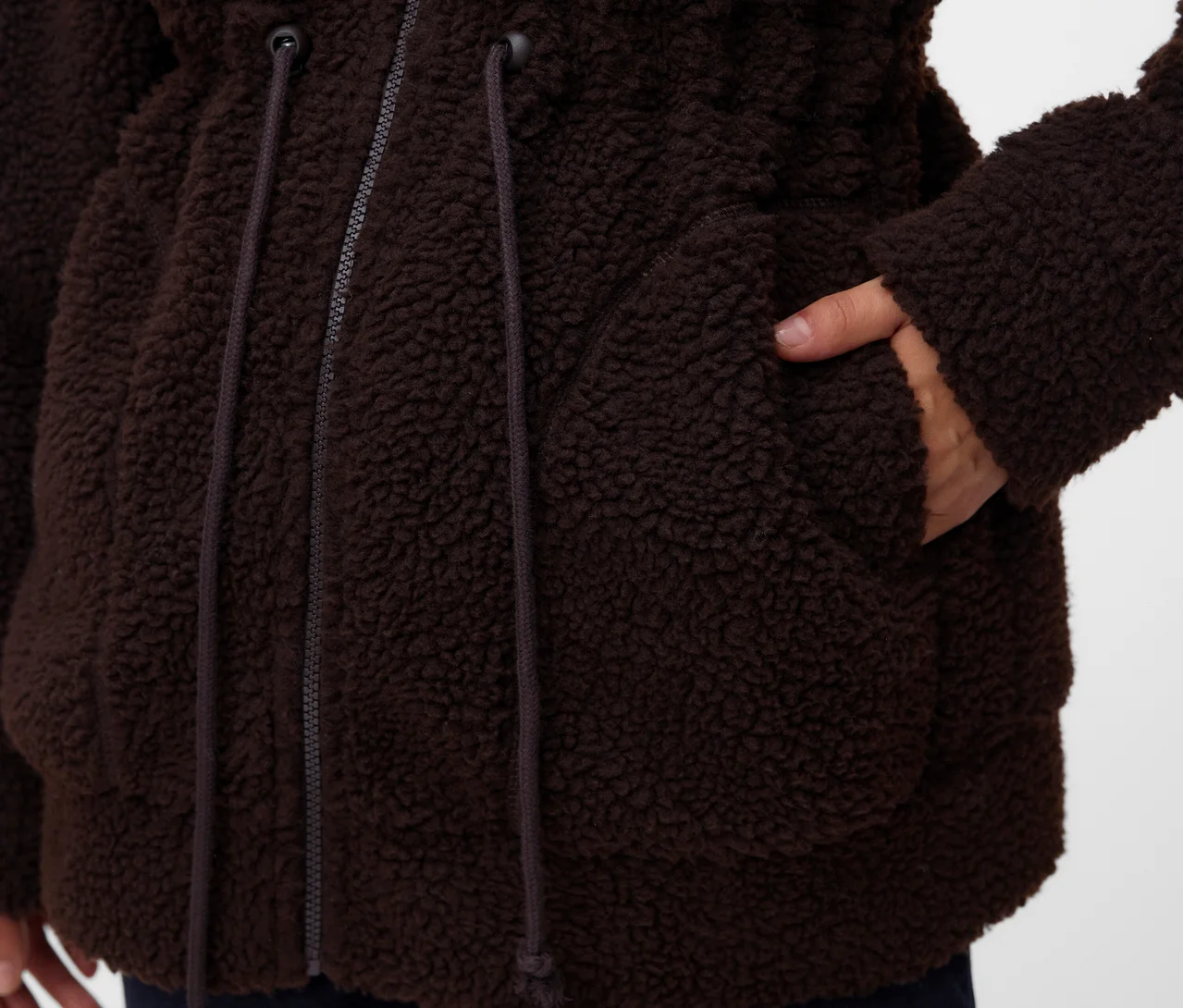 Detail osoby v hnědé, huňaté bundě s rukou v kapse. Bunda má zip a stahovací šňůrky v pase.