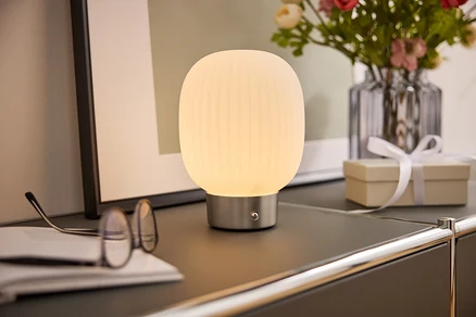 Noční lampa s bílým stínidlem stojí na skříňce vedle brýlí, knihy, vázy s květinami a dárkové krabičky.