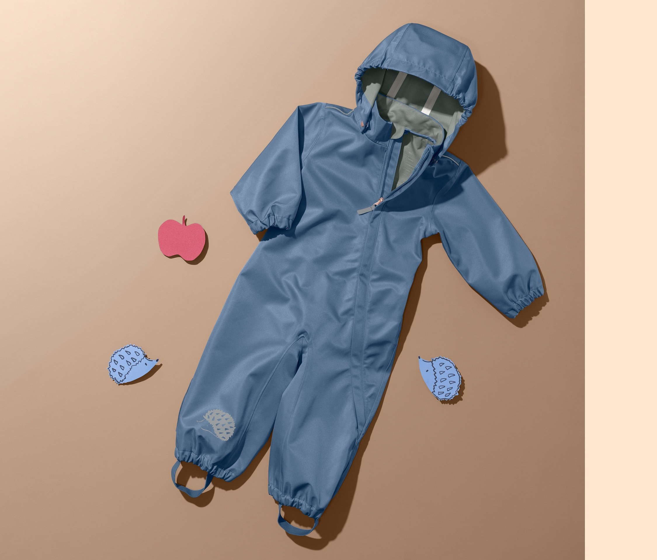 Modrá kombinéza do deště leží na hnědém pozadí vedle dvou ježků a jablka.