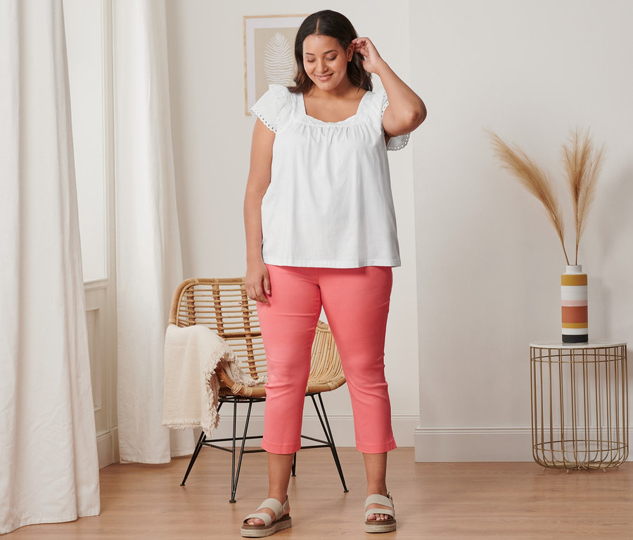 Žena má na sobě korálové 7/8 strečové kalhoty, bílé triko s výšivkou a kožené sandály na platformě.