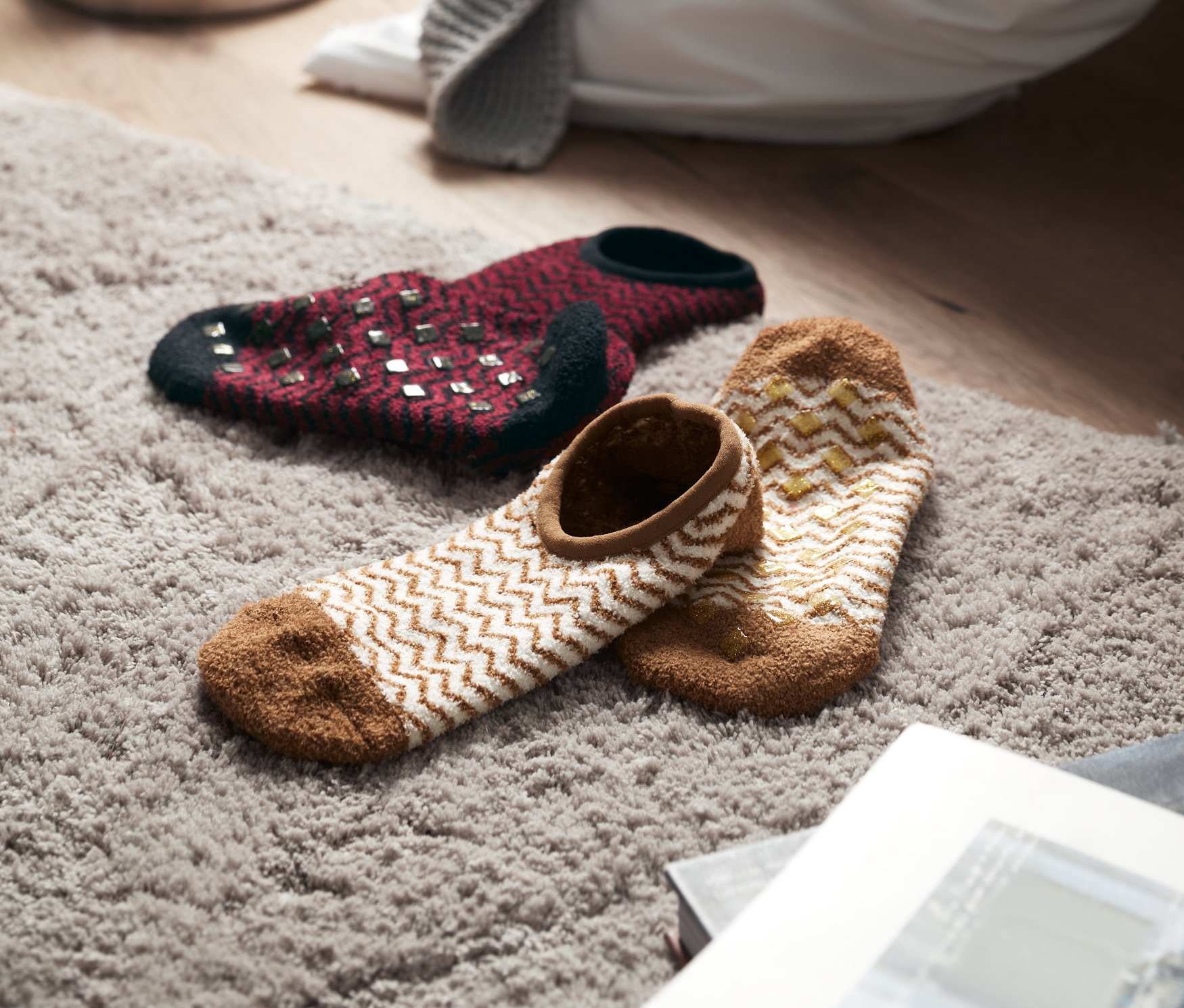 Dva páry ponožkových bačkor leží na šedém koberci, v pozadí svíčky a deka.