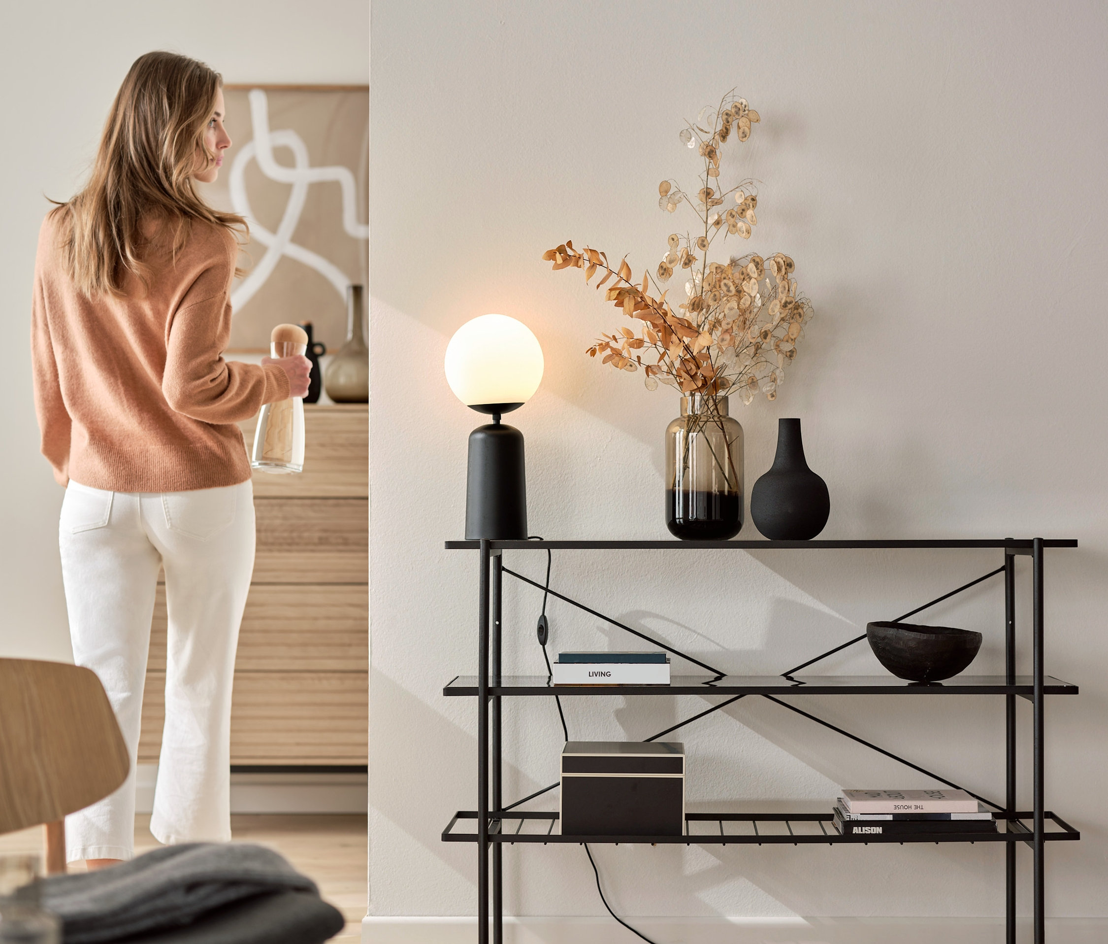 Žena drží láhev u konzolového stolku (šířka cca 120 cm) s černou stolní lampou se skleněnou koulí.