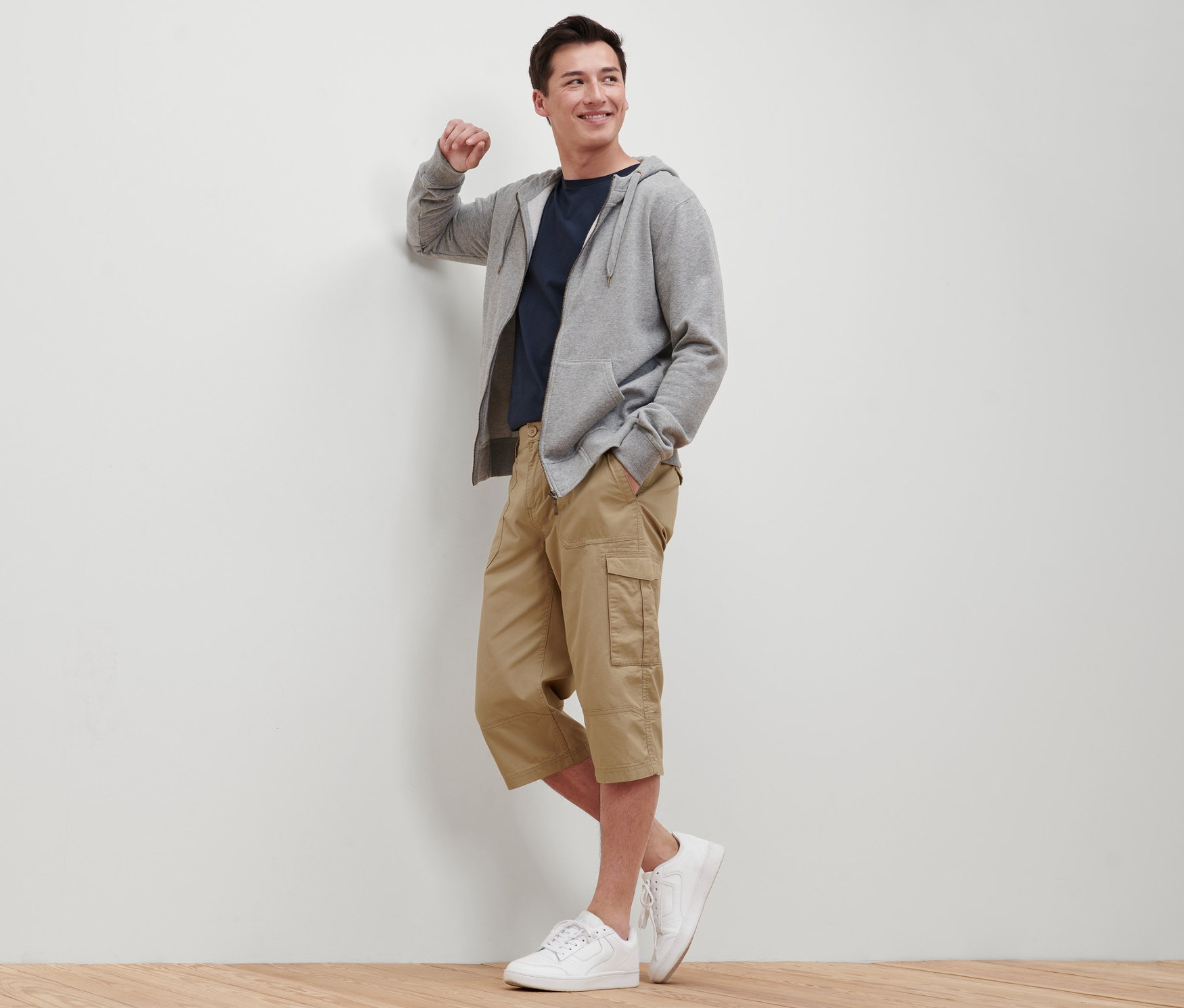 Muž opřený o zeď má na sobě béžové cargo bermudy ve 3/4 délce, tričko, mikinu s kapucí a tenisky.