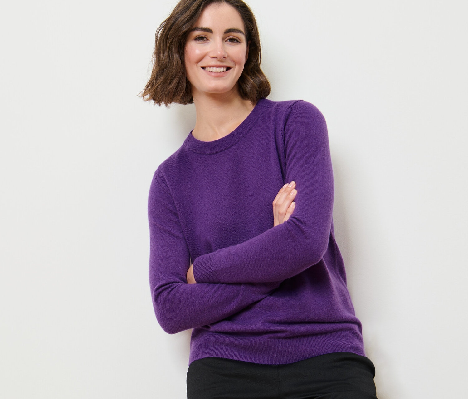 Žena se zkříženýma rukama pózuje v fialovém svetru z jemného úpletu s kašmírem a černých kalhotách.