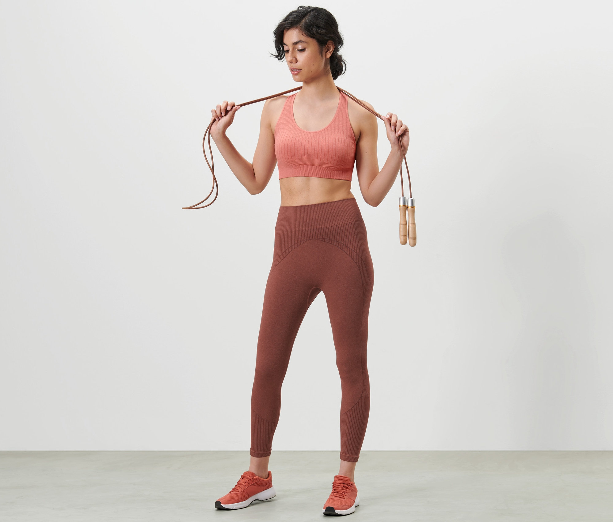 Žena drží švihadlo na ramenou, má na sobě sportovní podprsenku a 7/8 bezešvé funkční sportovní legíny.
