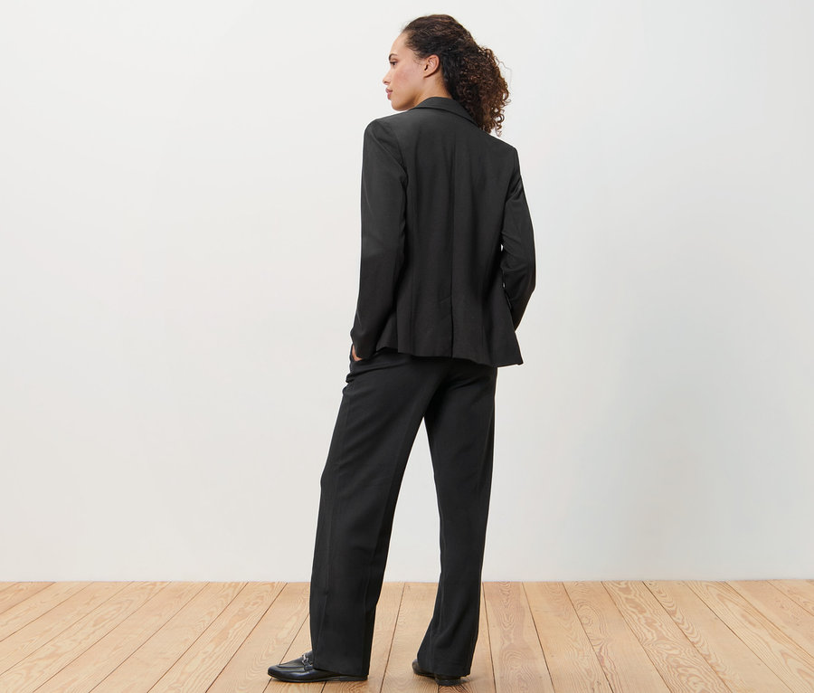 Žena v černém žerzejovém blejzru a kalhotách stojí zády ke kameře.