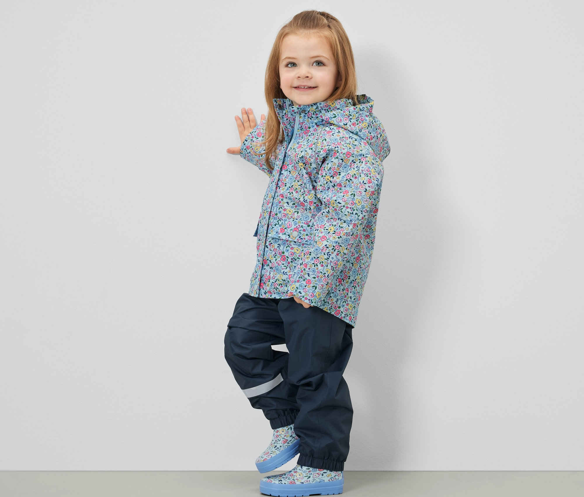 Dívka stojí bokem a má na sobě dětskou bundu do deště s květinovým vzorem, tmavě modré kalhoty a boty.