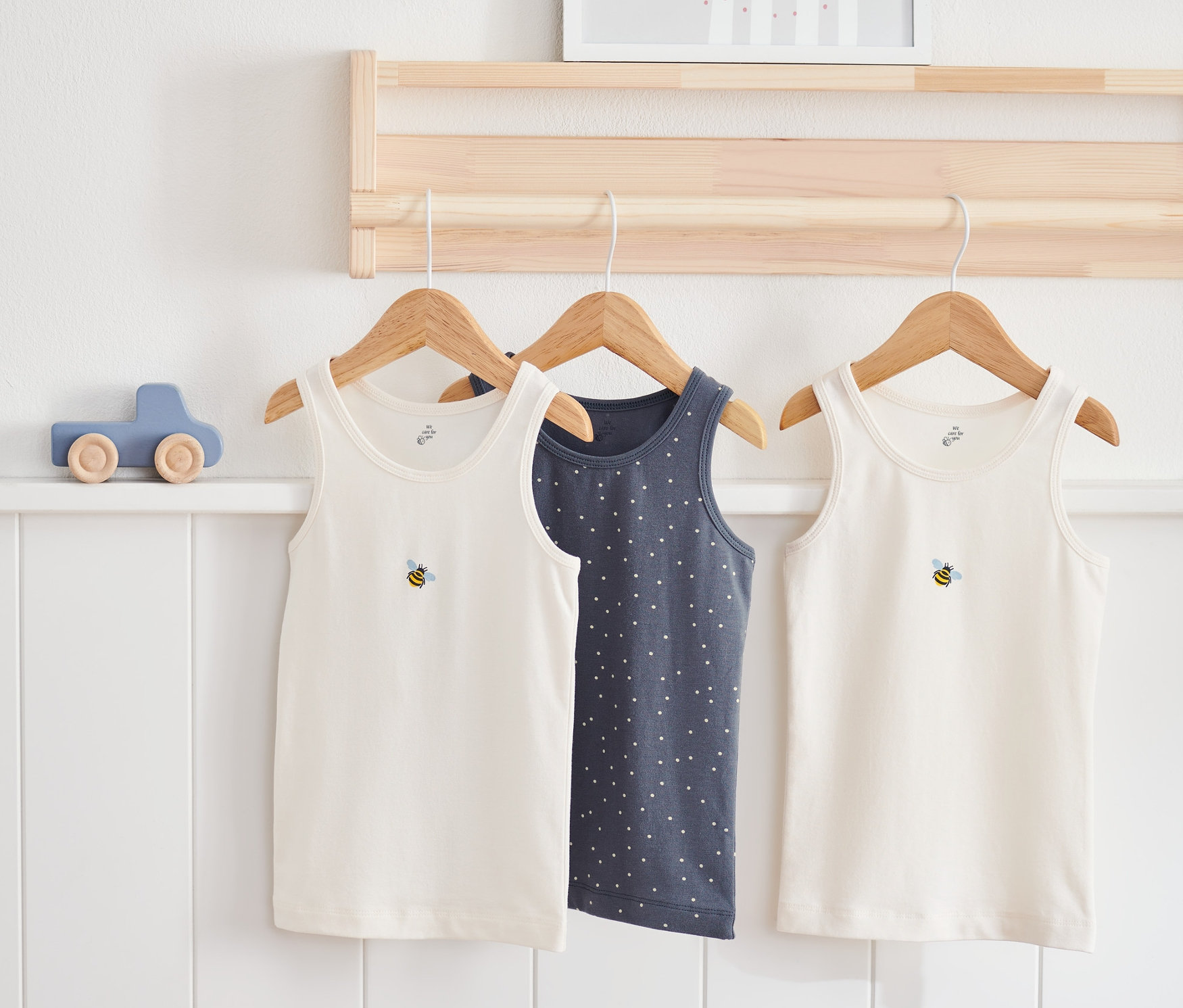 Tři dětské košilky visí na dřevěném věšáku. Dvě jsou bílé s motivem včely, jedna je tmavě modrá s bílými puntíky.
