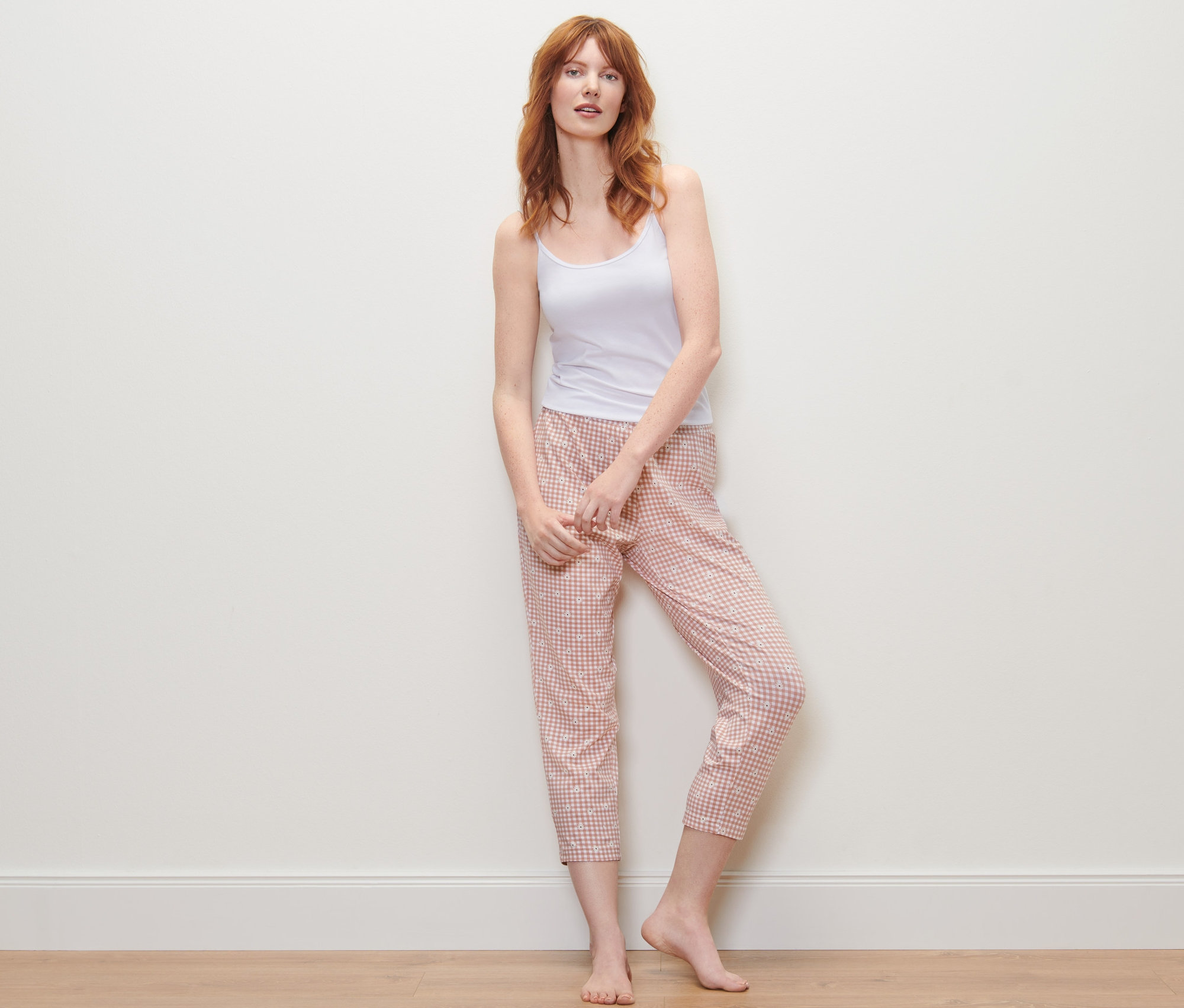 Červenovlasá žena stojí a má na sobě bílý top a kostkované 7/8 pyžamové kalhoty.